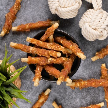 Eine schwarze Schüssel, gefüllt mit Hundeleckerlis in Form von Stäbchen, umhüllt von knusprigem Fleisch, umgeben von einem Seilspielzeug und einer kleinen Pflanze.