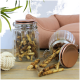 Ein Glasbehälter mit Hundekauartikeln steht auf einem Holztisch, einige Kauartikel sind herausgefallen. Im Hintergrund befindet sich ein weiches Haustierbett.