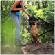 Eine Frau in einem schwarzen Tanktop und einer blauen Hüfttasche hält eine Hundeleine, steht neben einem sitzenden braunen Hund in einem grünen Wald.