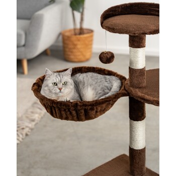 Eine graue Katze, die in einer gemütlichen braunen Hängematte eines Katzenbaums entspannt, mit einem hängenden Spielzeug darüber und einem modernen Wohnzimmer im Hintergrund.