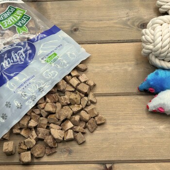 Verschüttete Tüte mit Würfel-Hundesnacks auf einem Holztisch neben einem Tauknoten und zwei Plüschmäusen, Verpackung mit der Aufschrift 'NATURE POWER'.