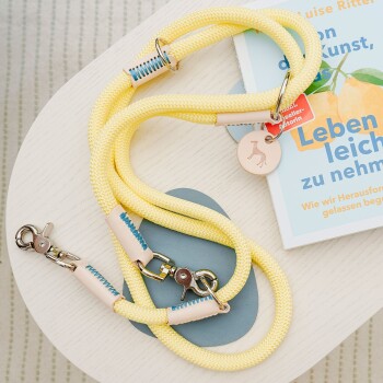 Eine leuchtend gelbe Hundeleine mit Lederakzenten und Metallclips, die auf einem hellen Holztisch neben einem Buch mit dem Titel 'Leben leicht zu nehmen' ruht.