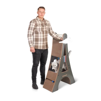 Design-Kratzmöbel Eiffelturm Eine Person steht neben einem stilvollen Kratzbaum für Katzen, der dem Eiffelturm ähnelt, mit einer grauen Katze, die aus einer der Ebenen herausspäht.