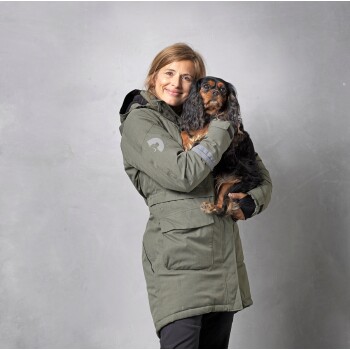 Eine Person in einer olivgrünen Jacke hält einen kleinen, flauschigen Hund nah an ihrer Brust, vor einem grauen Hintergrund.