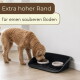Brauner, lockiger Hund frisst aus einem Doppelnapf in einem schwarzen Tablett; deutscher Text: 'Extra hoher Rand' und 'für einen sauberen Boden'.