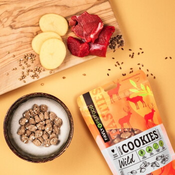 Ein glücklicher Hund sitzt in einem Wald neben einer bunten Verpackung von "FAVLY PET FOOD Cookies Wild", die hypoallergene, getreidefreie Zutaten hervorhebt.