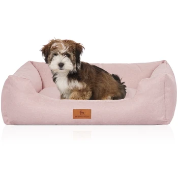 Hundebett Emma aus Velours mit feinem Handwebcharakter in Pastellfarben hellrosa M-L Ein kleiner, flauschiger Hund sitzt bequem in einem weichen, rosa Haustierbett mit einem braunen Lederetikett, auf dem "KNUFFELWUFF" steht.