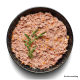 Eine dunkle Schüssel mit zerkleinertem Haustierfutter, garniert mit frischem Rucola und dem Text "Serviervorschlag" in der unteren rechten Ecke.