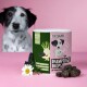 Eine Dose 'Darmpflege Drops' Haustierergänzungen auf einer rosa Oberfläche, mit Kräutern, einer Gänseblümchen und einem schwarz-weißen Hund im Hintergrund.