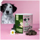 Eine Dose 'Darmpflege Drops' Haustierergänzungen auf einer rosa Oberfläche, mit Kräutern, einer Gänseblümchen und einem schwarz-weißen Hund im Hintergrund.