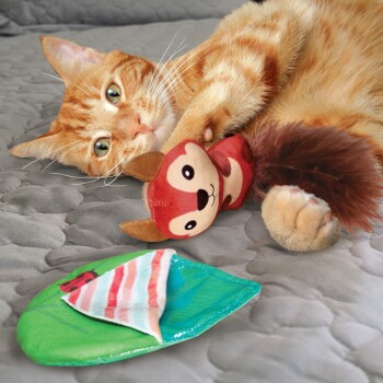Eine orange getigerte Katze spielt mit einem plüschigen Eichhörnchen-Spielzeug neben einem grünen fischförmigen Spielzeug auf einer grauen, gesteppten Oberfläche.