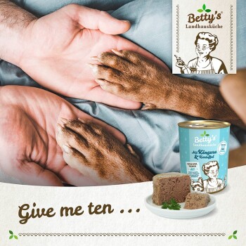 Eine Hand und die Pfote eines Hundes berühren sich, mit Betty's Landhausküche Känguru-Hundefutter und einem Teller im Hintergrund. Text: "Gib mir zehn ...".