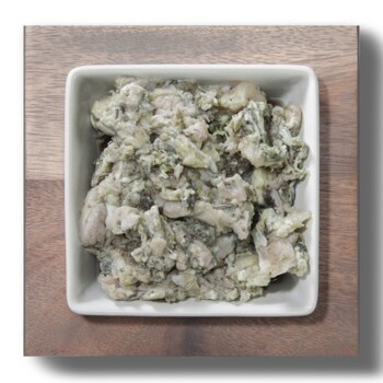 Eine weiße Schüssel, gefüllt mit einer stückigen, grünlich-grauen Haustierfuttermischung, auf einer Holzoberfläche platziert.