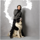 Eine Person in einem grauen Hoodie und schwarzen Hosen steht neben einem großen, flauschigen schwarz-weißen Hund, beide vor einem grauen Hintergrund.