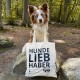 Ein Hund steht in einem Wald, mit dem Rücken zur Kamera, und hält eine Tragetasche mit dem Text "HUNDE LIEB HABER", der deutlich sichtbar ist.