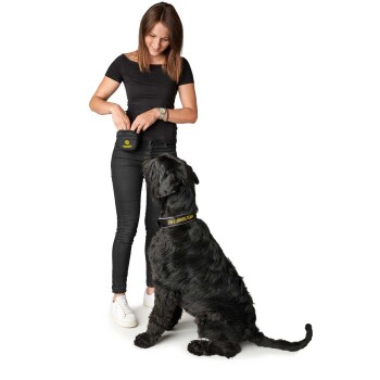 Eine Frau in einem schwarzen Shirt und Jeans hält eine Leckerlitasche, während ein großer schwarzer Hund mit einem Halsband, das "EIN SANKTES TEAM" beschriftet ist, vor ihr sitzt.