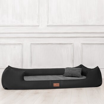 Schwarz-graues rechteckiges Haustierbett mit erhöhten Seiten und kleinem Kissen, auf hellem Holzboden platziert.