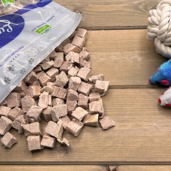 Verschüttete Tüte mit würfelförmigen Haustierleckerlis auf einem Holztisch, rechts Seil- und Plüschspielzeug; auf der Tüte steht '100%'.