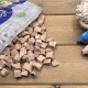 Verschüttete Tüte mit würfelförmigen Haustierleckerlis auf einem Holztisch, rechts Seil- und Plüschspielzeug; auf der Tüte steht '100%'.