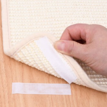 Eine Hand hält den Rand einer cremefarbenen Haustiermatte und zeigt einen Klettverschlussstreifen darunter, mit einem kleinen Klettstück daneben auf einer Holzoberfläche.