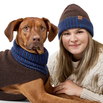 Ein Hund, der einen braunen Pullover mit blauen Streifen trägt, sitzt neben einer Frau in einem cremefarbenen, kabelgestrickten Pullover, beide sehen gemütlich aus.