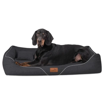 WILLIAM Couture - Orthopädisches Hundebett anthrazit 1,1 m, 22 cm, 85 cm Ein schwarz-brauner Hund, der bequem auf einem dunkelgrauen Hundebett mit weißer Paspelierung liegt, das ein kleines Kissen und ein Markenetikett hat.
