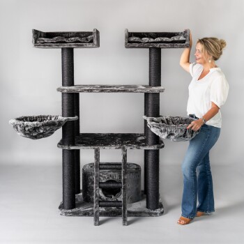 Eine Frau in einer weißen Bluse und blauen Jeans montiert einen großen, mehrstöckigen Katzenbaum mit plüschigem grauem Stoff und hängenden Körben.