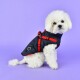 Ein flauschiger weißer Hund, der eine marineblaue und rote Haustierjacke trägt, sitzt vor einem lila Hintergrund und schaut neugierig in die Kamera.