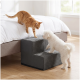 Weißer, flauschiger kleiner Hund steigt eine graue Haustiertreppe mit zwei Stufen hinauf, während eine orangegetigerte Katze vom Bett hinunterreicht in einem hellen Schlafzimmer.