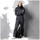 Eine Person in einem schwarzen isolierten Overall steht selbstbewusst mit verschränkten Armen neben einem schwarzen Hund, vor einem grauen Hintergrund.