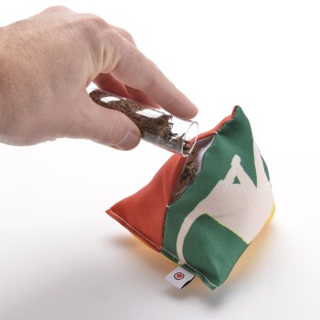 Eine Hand, die getrocknete Kräuter aus einem Glasgefäß in ein buntes, dreieckiges Haustierspielzeug mit einem Katzendesign gießt.