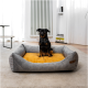 Ein kleiner schwarz-brauner Hund sitzt auf einem gemütlichen grauen Hundebett mit einem gelben Kissen in einem hellen Raum mit weißen Vorhängen und einem Heizkörper.
