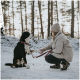 Eine Frau in einem gemütlichen Outfit kniet im Schnee, hält eine Leine, während ihr Berner Sennenhund ihr die Pfote anbietet, mit Kiefern im Hintergrund.