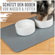 Ein Hund trinkt aus einer weißen Schüssel auf einer grauen Matte, mit dem Text "SCHÜTZT DEN BODEN VOR WASSER & FUTTER" darüber.