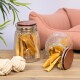 Ein Glasbehälter gefüllt mit Hundeleckerlis, einige fallen auf einen Holztisch, neben zwei weißen Seilspielzeugen und einem rosa Haustierbett im Hintergrund.