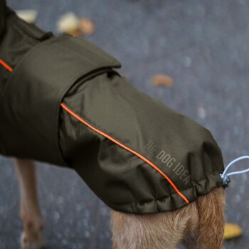 Ein Hund trägt einen braunen wasserdichten Mantel mit orangefarbenen Akzenten, der den Text "DIE HUND IDEE" an der Seite zeigt und auf einem Gehweg steht.