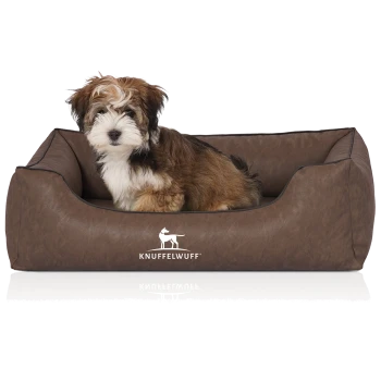 Ein kleiner, flauschiger Hund sitzt bequem in einem braunen Haustierbett mit dem Logo "KNUFFELWUFF", das prominent auf der Vorderseite angezeigt wird.
