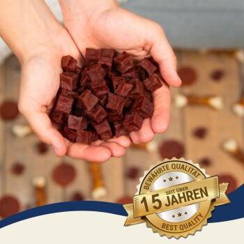 Hände halten einen Haufen kleiner, dunkelroter Haustierleckerlis, mit einem Abzeichen, das 'Beste Qualität' und '15 Jahre' auf Deutsch angibt.