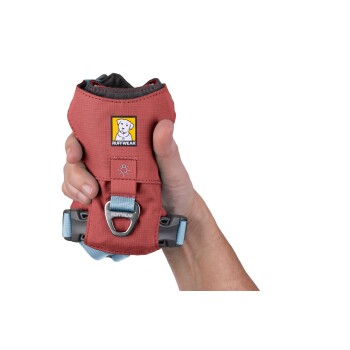 Eine Person hält ein rotes Hundegeschirr mit einem blauen Riemen und einem Metallclip, das das Ruffwear-Logo auf der Vorderseite zeigt.