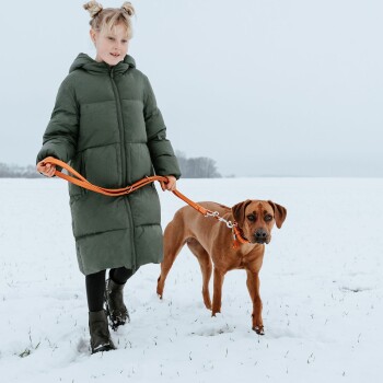 Eine Person in einer grünen Daunenjacke führt einen braunen Hund an einer orangefarbenen Leine durch ein verschneites Feld unter einem grauen Himmel.
