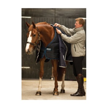 Eine Person stattet ein Pferd mit einer blauen und schwarzen Pferde-Decke in einem Stall aus. Das Pferd hat ein braunes Fell und eine weiße Blesse.