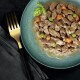 Gros plan d'une assiette teal remplie de nourriture humide pour animaux, avec des morceaux de viande, des petits pois verts et des carottes en dés, garnie d'une feuille de basilic. Une fourchette dorée repose à côté d'un tissu sombre.