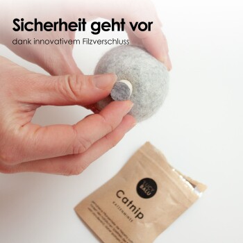 Eine Person hält einen grauen Filz-Katzenspielball mit einem Sicherheitsverschluss, während ein teilweise geöffnetes Paket mit der Aufschrift 'Catnip' in der Nähe sichtbar ist.