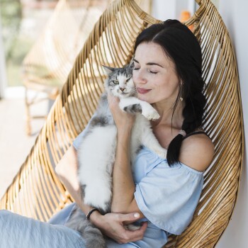 Eine Frau in einem hellblauen schulterfreien Kleid hält eine flauschige grau-weiße Katze, während sie in einem geflochtenen Stuhl sitzt.