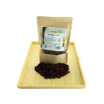 Ein Paket mit getrockneten roten Rübenflocken auf einem Holztablett, mit verstreuten Flocken und einem Etikett mit der Aufschrift "Rote Beetflocken, 100% frisch."