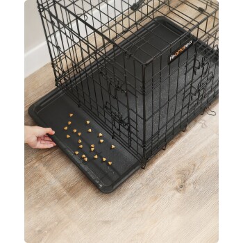 Eine Hand, die Hundeleckerlis auf das schwarze Kunststofftablett eines Draht-Haustierkäfigs legt, wobei der Käfig teilweise geöffnet ist und ein "Feandrea"-Logo sichtbar ist.