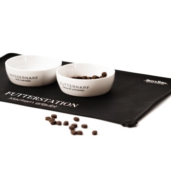 Zwei weiße Fressnäpfe mit der Aufschrift "WASSERNAPF" und "FUTTERNAPF" auf einer schwarzen Matte, mit verstreutem Trockenfutter in der Nähe.