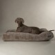 Ein grauer Weimaraner-Hund liegt auf einem plüschigen, rechteckigen Hundebett mit gestepptem Design und schaut neugierig in die Kamera.