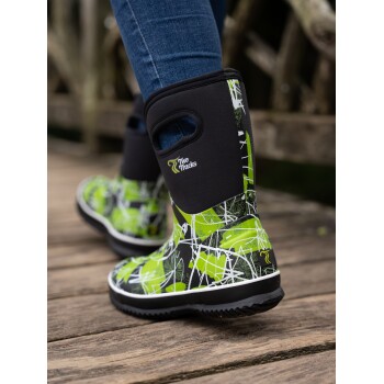 Nahaufnahme der Beine einer Person, die lebhaft grüne und schwarze gemusterte wasserdichte Stiefel mit dem Logo "Two Tracks" trägt, auf Holzplanken stehend.