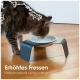 Eine hellgraue Katze sitzt neben einer modernen Futterschüssel auf einem Holzboden, mit einer großen grünen Pflanze dahinter. Text: "Stabil & sicher für stressfreies Essen."